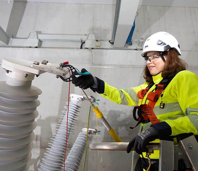 Technician in high-visibility gear working at dead tank high voltage circuit breaker, in a secure work zone.