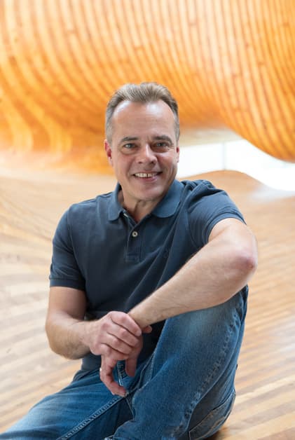 Andreas Claus in a grey shirt sitting on a wooden sculpture, representing Innovation Process Management with a statement.