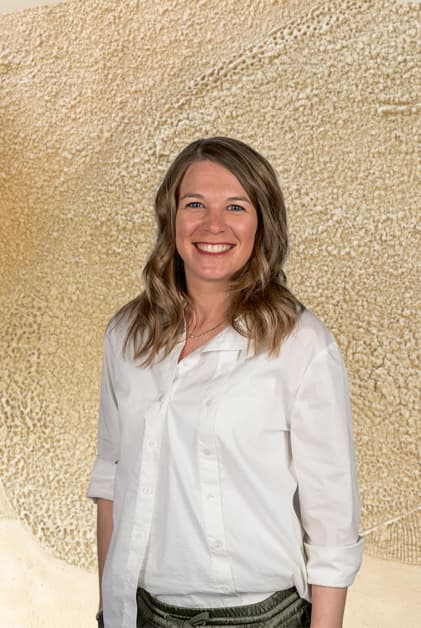 Jennifer Lebeda in a white shirt standing in front of a neutral wall, representing OMICRON Academy with a statement.