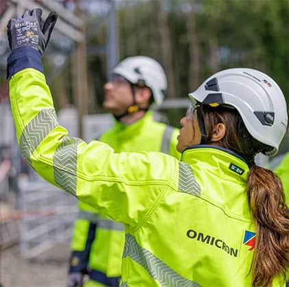 OMICRON field technician raising a gloved hand while working in an substation, accompanied by a colleague in safety gear.