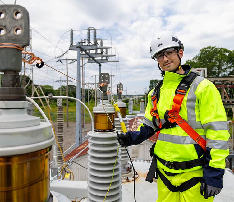 Technician in high-visibility gear working at a high voltage power transformer, ensuring safe and reliable power infrastructure operations.