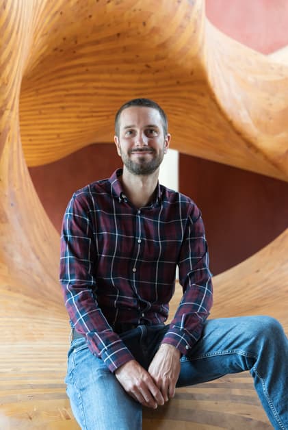 Simon Lecker in a red shirt sitting on a wooden sculpture, representing Hardware Development with a statement.