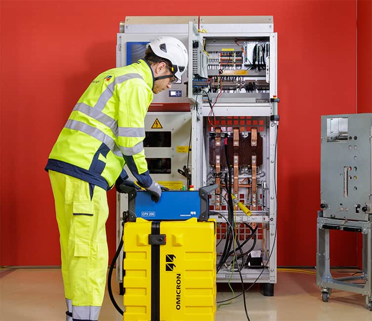 Technician in high-visibility gear using OMICRON CPX 200, performing diagnostics on an medium voltage instrument transformer, filled with circuit boards and wiring.