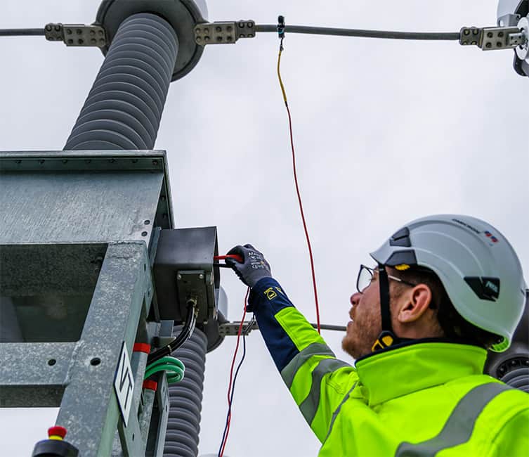 Technician in high-visibility gear working at a high voltage power transformer, ensuring safe and reliable power infrastructure operations.