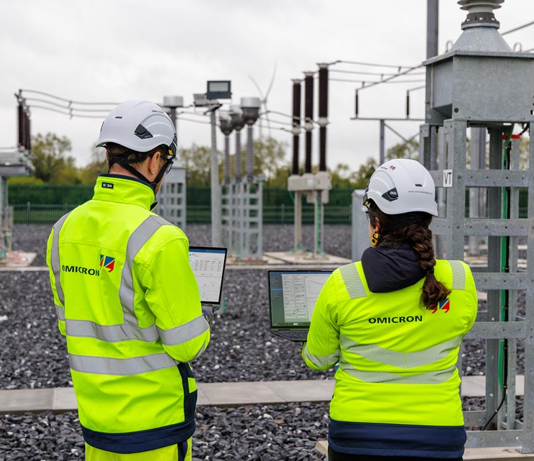 Two OMICRON engineers in safety gear using laptops at a power substation with a data management software, surrounded by electrical infrastructure.